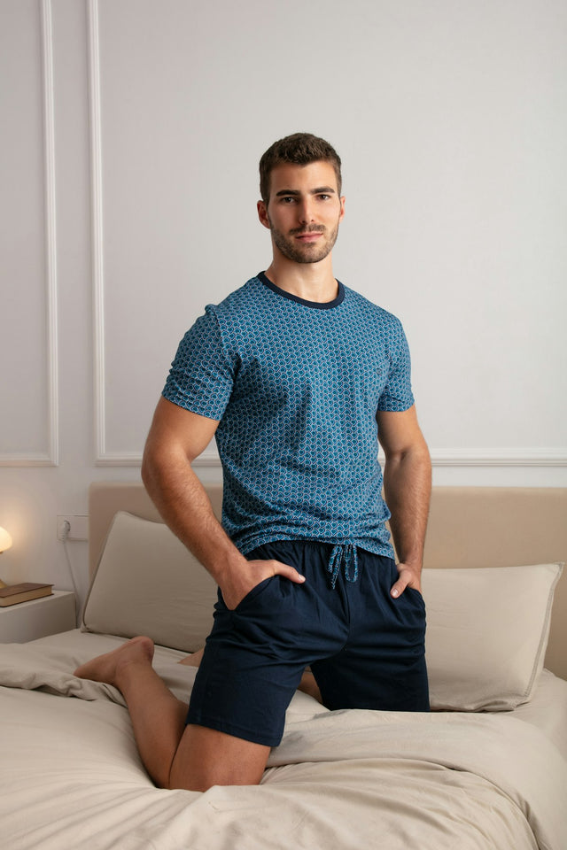 Man wearing a blue patterned shirt and dark shorts sitting on a bed.