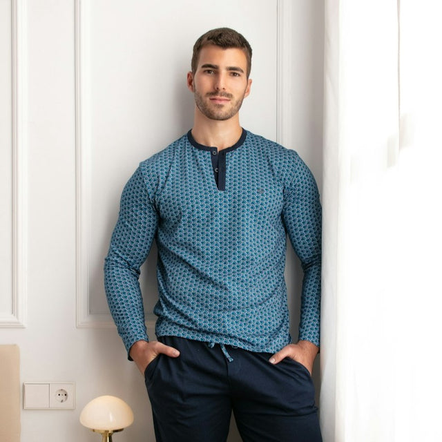 Man wearing a blue patterned long-sleeve shirt and dark pants against a white wall.