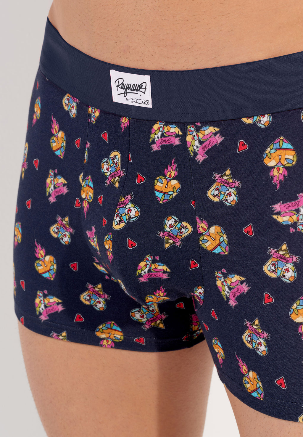male model wearing a pair of navy blue boxer briefs with a multicolored heart pattern and a wide red elastic waistband.