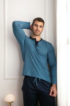 Man wearing a blue patterned shirt and dark pants against a white wall.