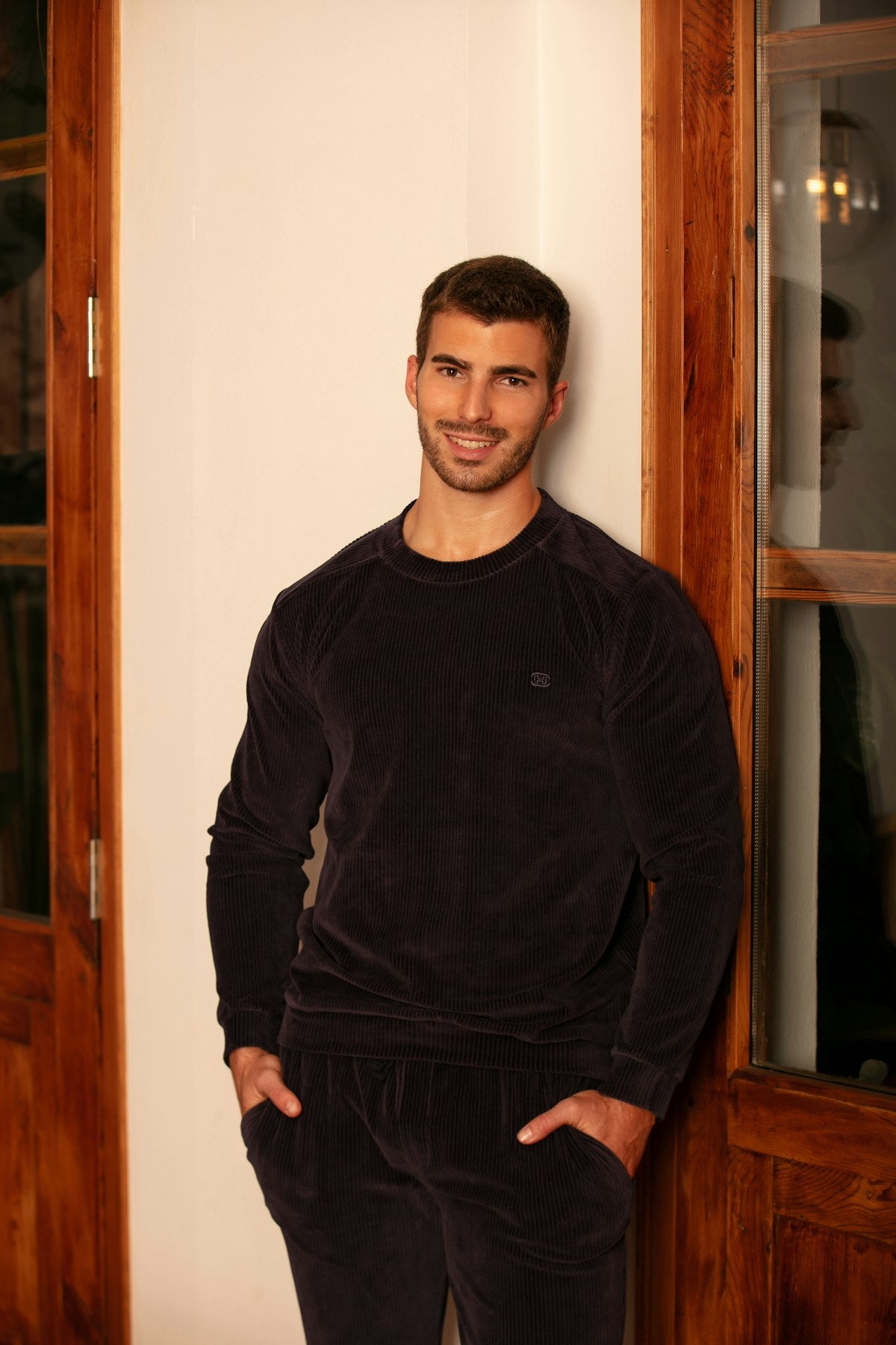 Man wearing a dark navy sweatshirt and pants standing against a wooden door.