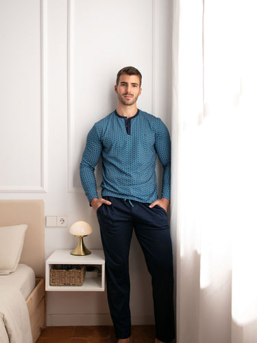 Man wearing a blue patterned shirt and dark pants standing in a bedroom.