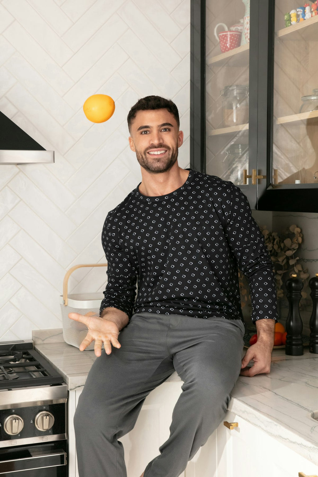 A man wearing a black long-sleeve crewneck shirt with a small white geometric pattern and gray pants, sitting on a kitchen counter while balancing an orange ball on his head.