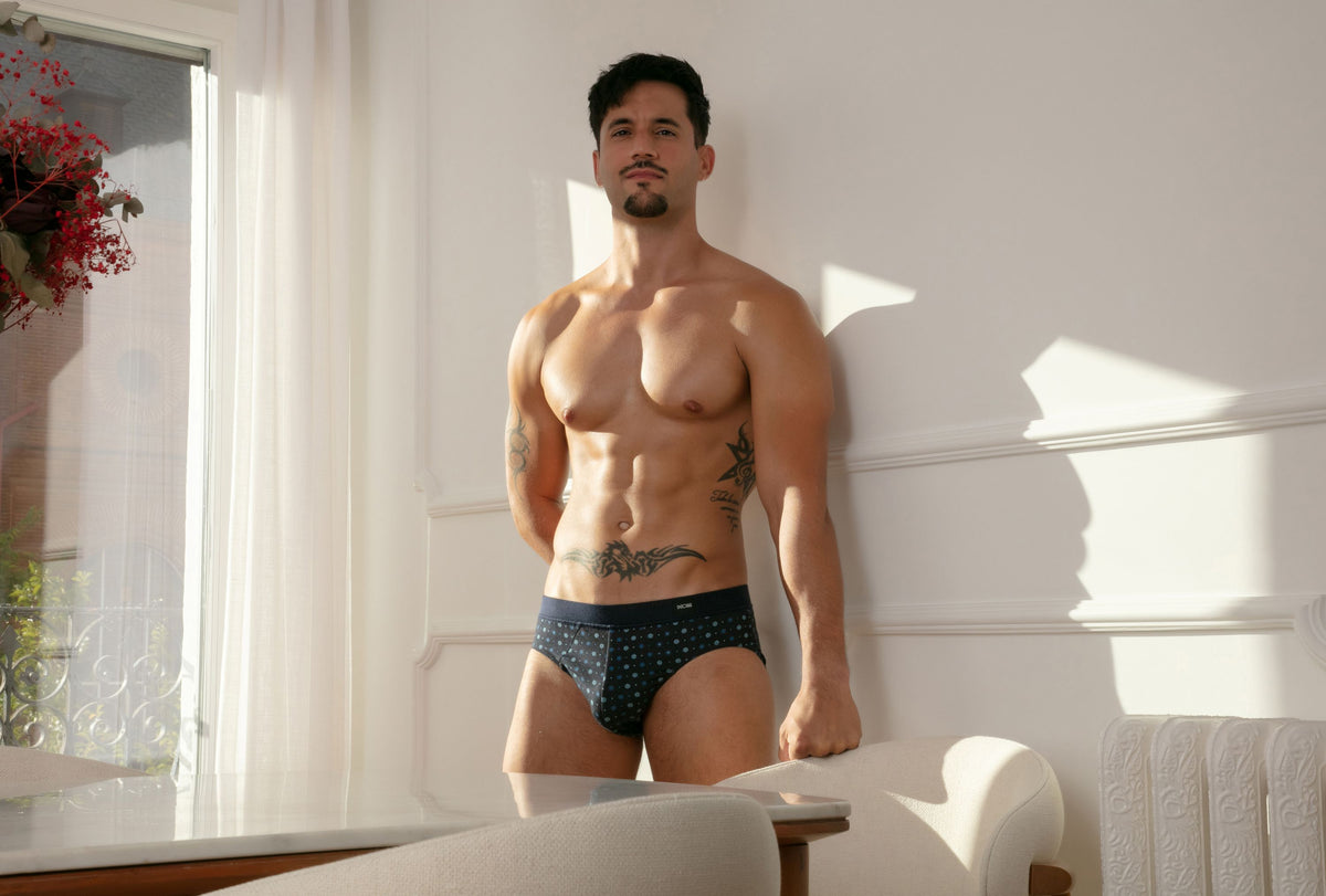 A muscular man with tattoos leans against a white wall in a brightly lit room, wearing dark navy patterned briefs.