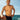 A shirtless man posing outdoors by the ocean, wearing black swim trunks under a blue sky.
