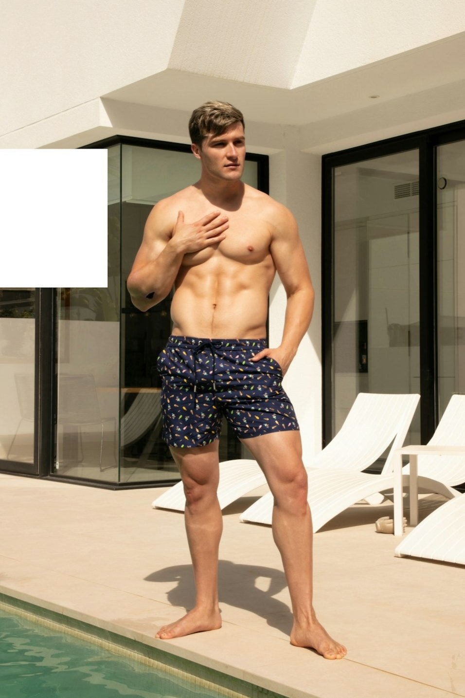 Man standing by a pool wearing navy swim trunks with white patterns.