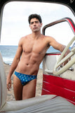 Man in a red, white, and blue swimsuit standing inside an open car on a beach.