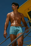 Man in blue swim trunks standing on a yellow lifeguard tower against a clear blue sky.