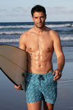 man wearing patterned swim shorts holding a surf board on the beach