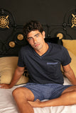 Man wearing a navy blue shirt and patterned shorts sitting on a bed with a decorative headboard.