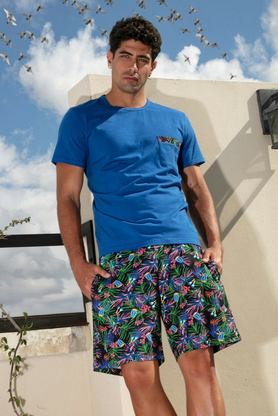 Man wearing a blue t-shirt and colorful shorts standing on a rooftop with birds flying around.