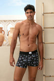 Man wearing black floral-patterned boxer briefs standing on a balcony with vases and a wall light fixture.