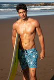 Man walking on a beach holding a surfboard, wearing blue patterned swim shorts.
