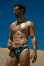 Man wearing a colorful striped swimsuit against a blue sky and sand dunes.