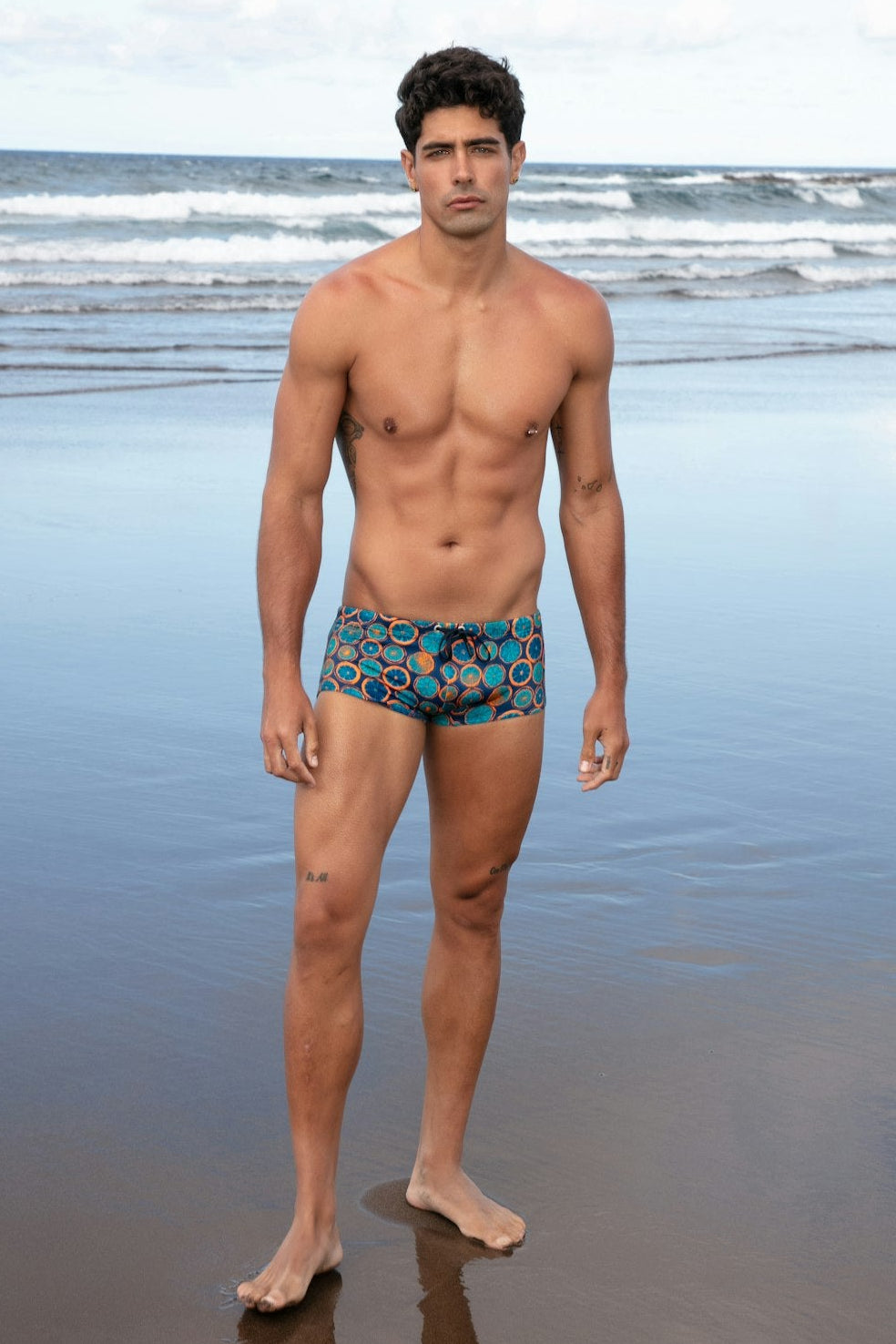 Man standing on a beach wearing patterned swim trunks with ocean waves in the background