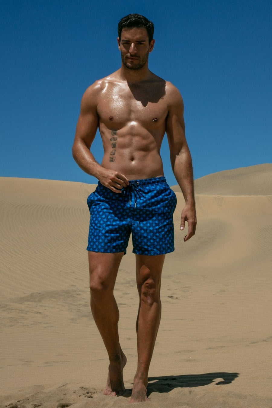 Man wearing blue swim shorts standing on sand with a clear blue sky