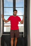 Man wearing a red t-shirt and patterned shorts standing by an open window with a blue sky view.
