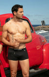Man standing shirtless next to a red vintage truck on a beach.