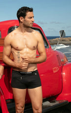 Man standing shirtless next to a red vintage truck on a beach.