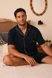 Man sitting on a bed wearing navy pajamas in a bedroom setting.