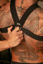 Person with tattoos holding a black harness over their chest.