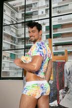 Man wearing a colorful tie-dye outfit standing in front of a window with an urban apartment complex view.