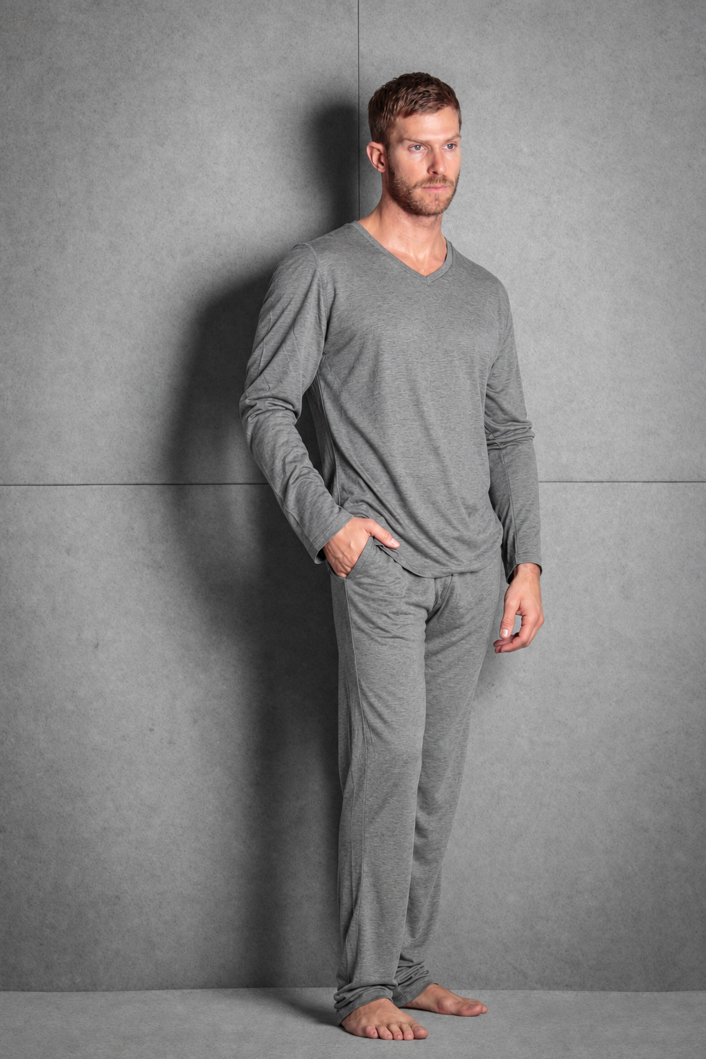 A man wearing a gray long-sleeve V-neck pajama shirt and matching gray pants, standing barefoot against a textured gray wall.